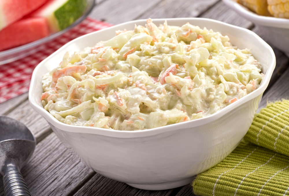 A bowl of delicious creamy homemade coleslaw on a rustic picnic table with watermelon and corn