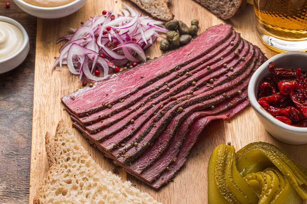 A wooden cutting board topped with sliced meat , pickles , onions , capers and a glass of beer.