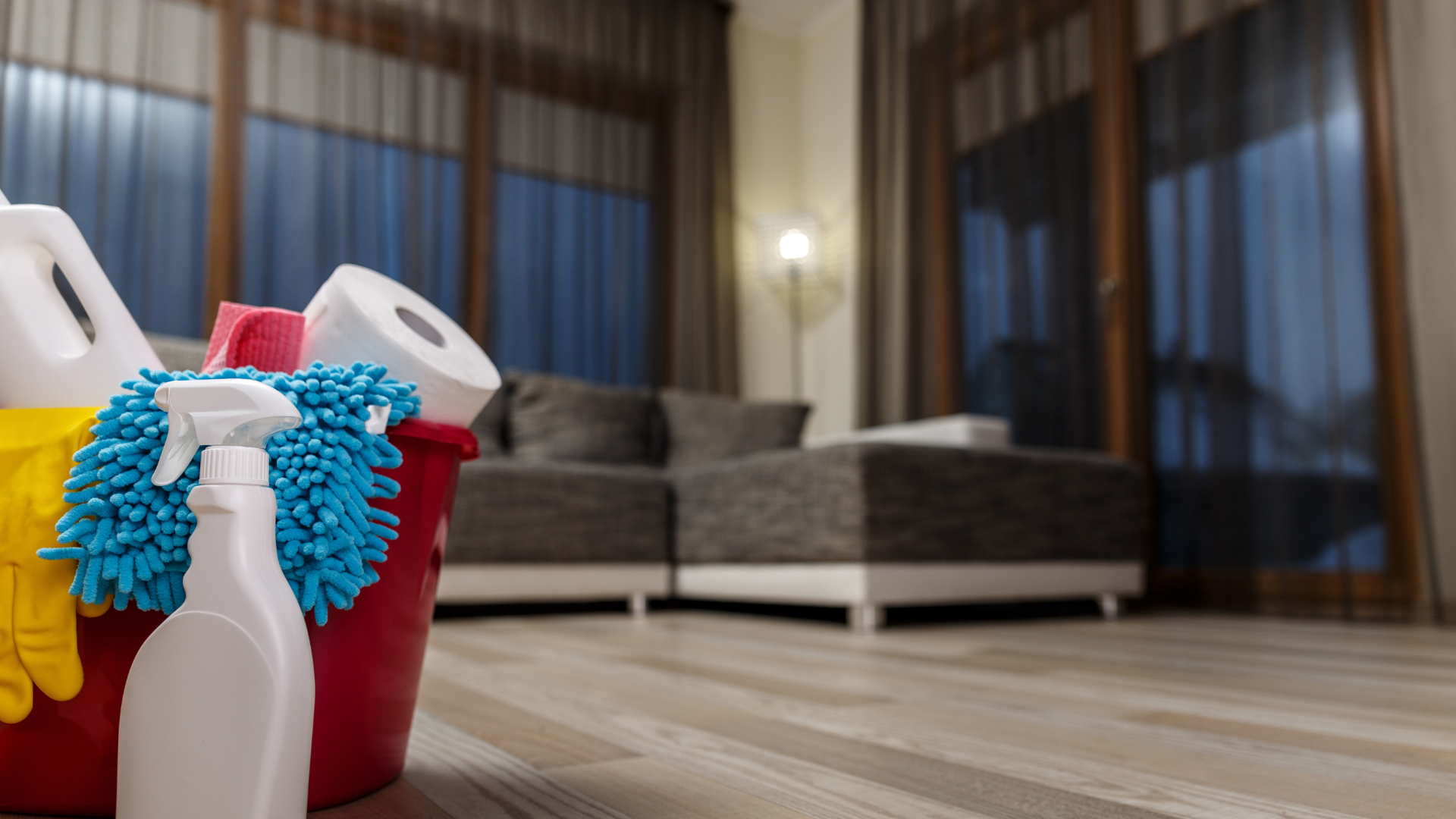 Cleaning supplies in a red bucket on a floor, with a couch and curtained windows in the background.