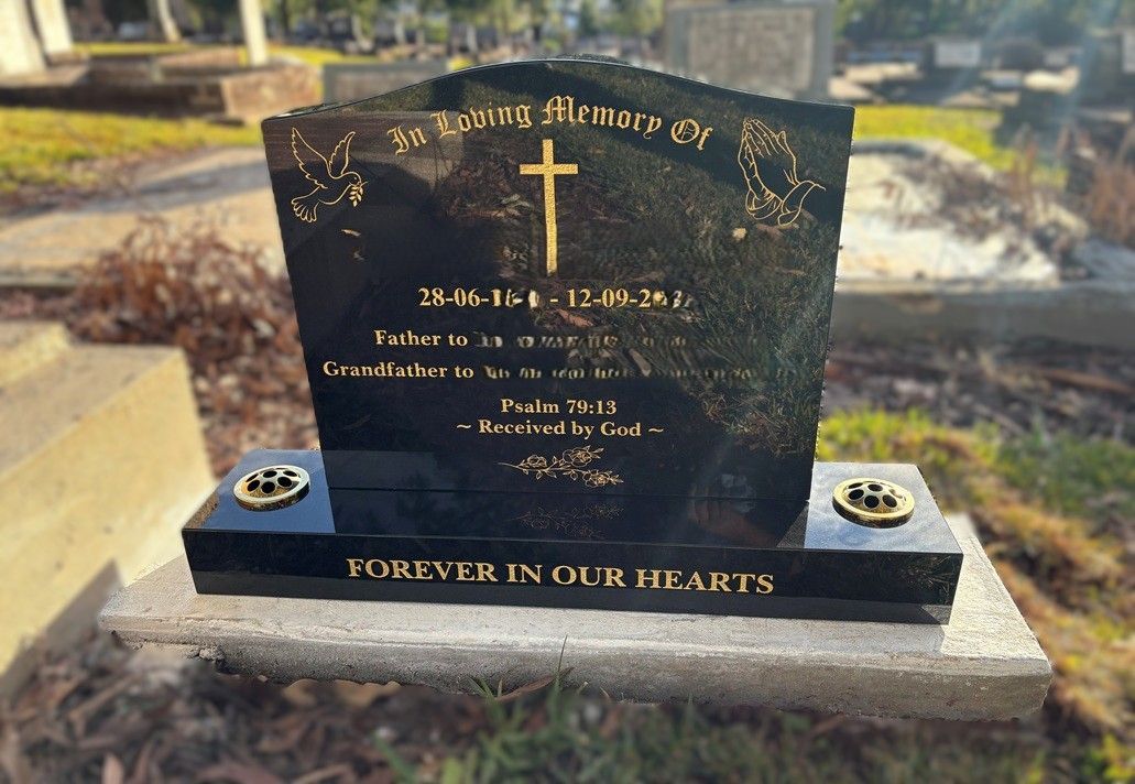 A black granite headstone with gold lettering and a cross, reading