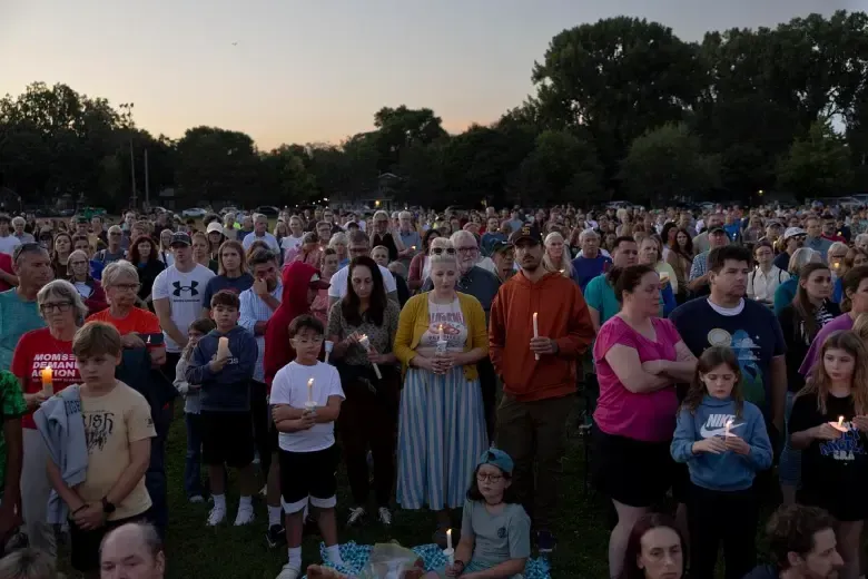 Large crowd at a vigil holding candles in a park at dusk. People are of diverse ages, expressions are somber.