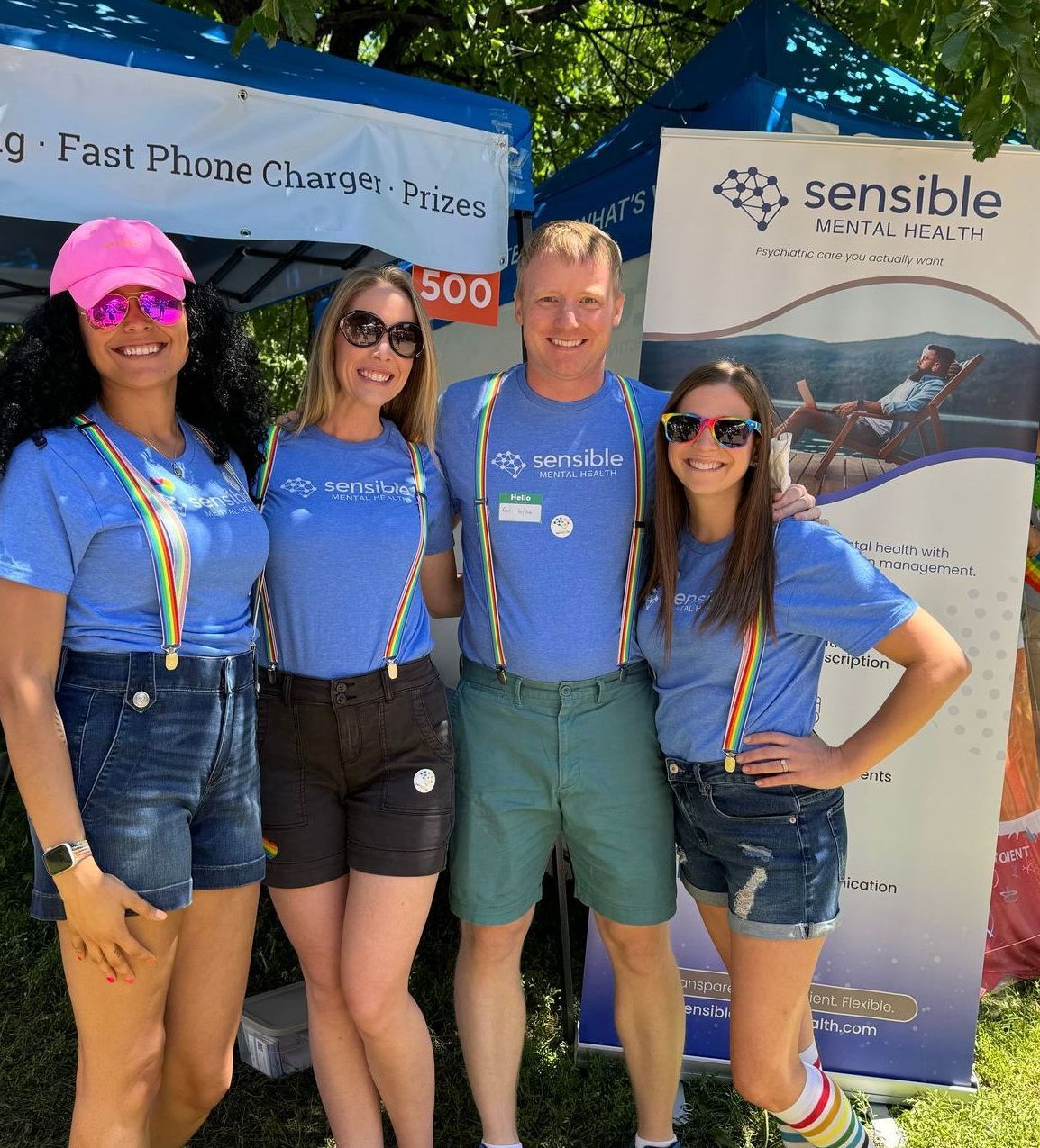Four people with rainbow suspenders pose in front of a booth. They wear blue shirts, denim shorts, and sunglasses.