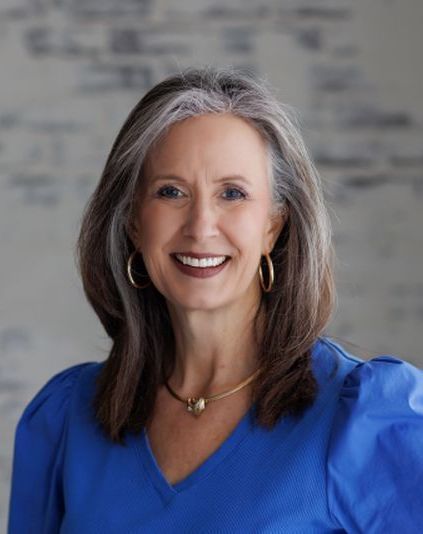 Woman with graying hair smiles, wearing a blue top and gold jewelry, against a neutral backdrop.