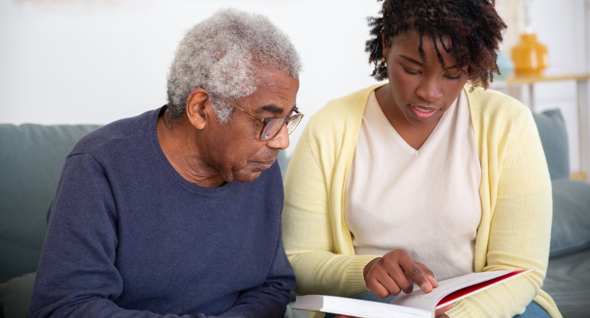 An elderly man and a young woman looking at a book together on a couch; the woman points at the book.