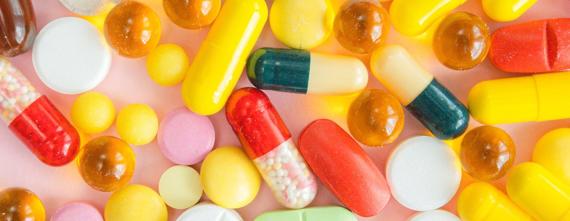 Assorted pills and capsules scattered on a light pink background. Various shapes and colors.