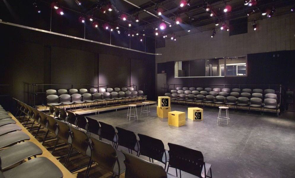 Empty black box theater with rows of black chairs facing the stage. Stage has three yellow cubes and a stool.