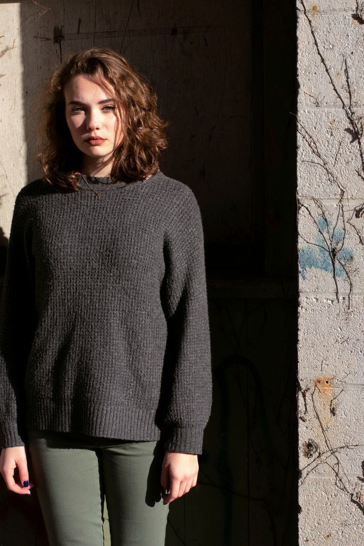 Woman in gray sweater and green pants, standing in sunlight next to a concrete wall with vines.