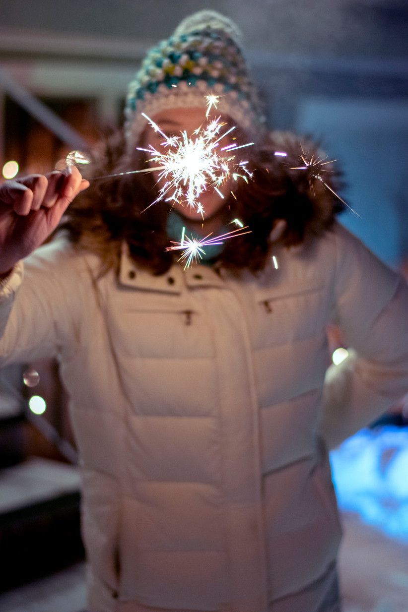 Person holding a sparkler, its light obscuring their face. They are wearing a white coat and hat, outdoors at night.