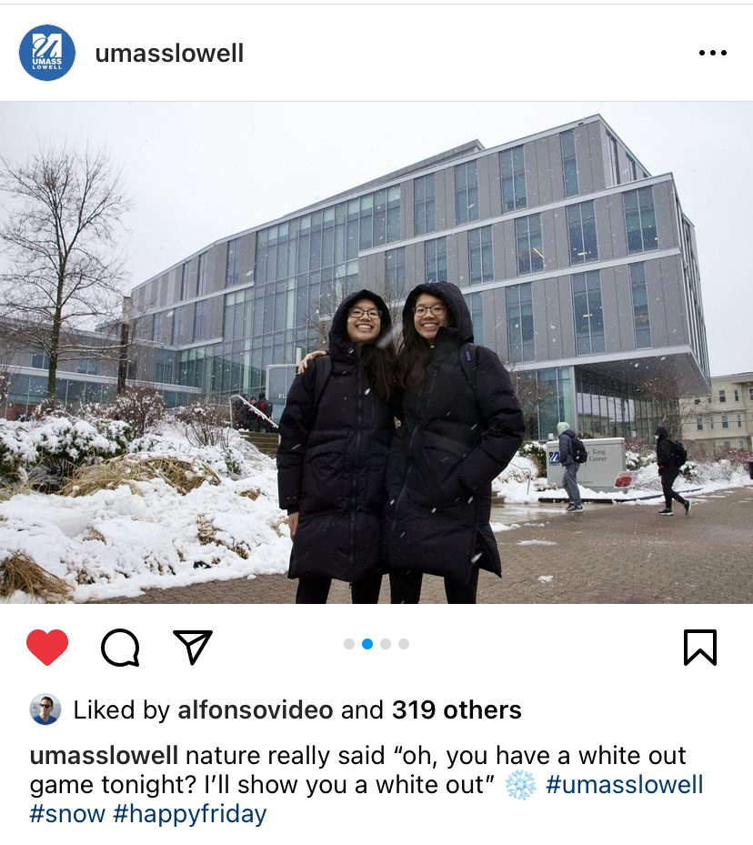 Two students in coats pose in front of a snow-covered building, UMass Lowell.