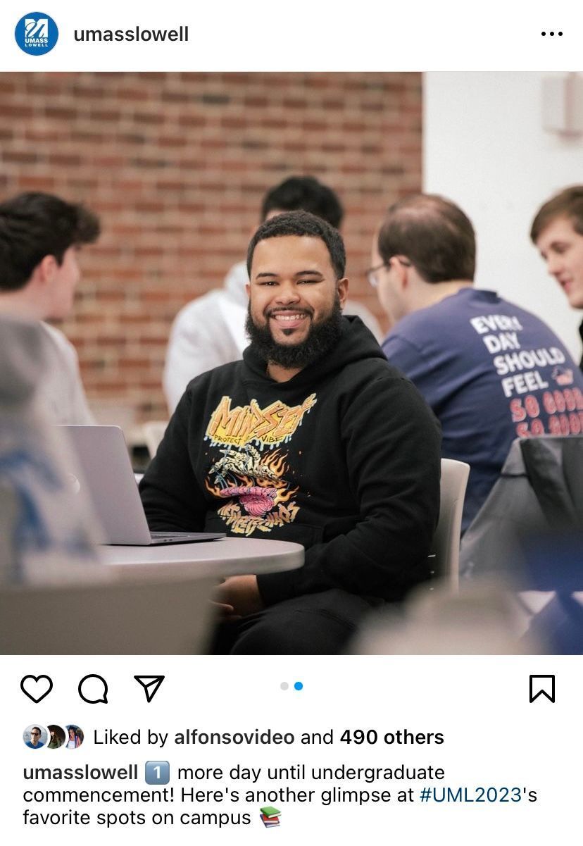 Man smiling, sitting at a table with a laptop, in a room with other students.