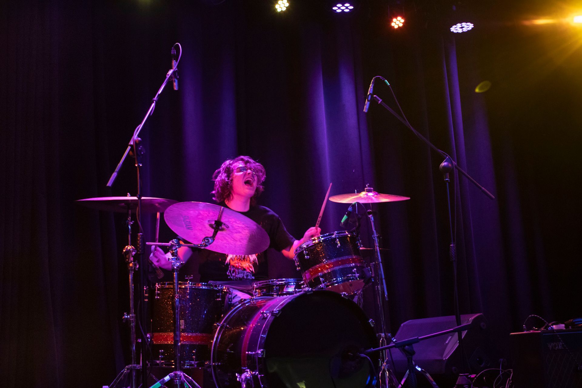 Drummer playing a drum kit on a stage with purple and yellow stage lighting.