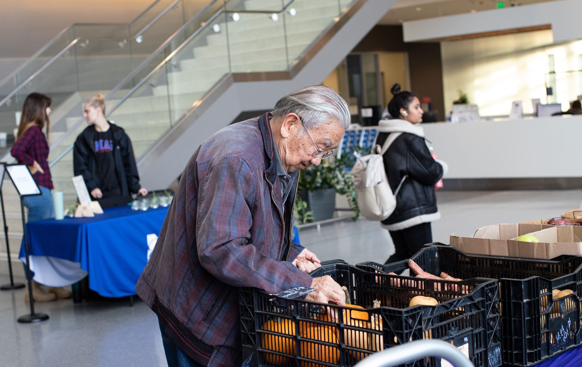 Older person selecting produce from crates at an event table, others in background.