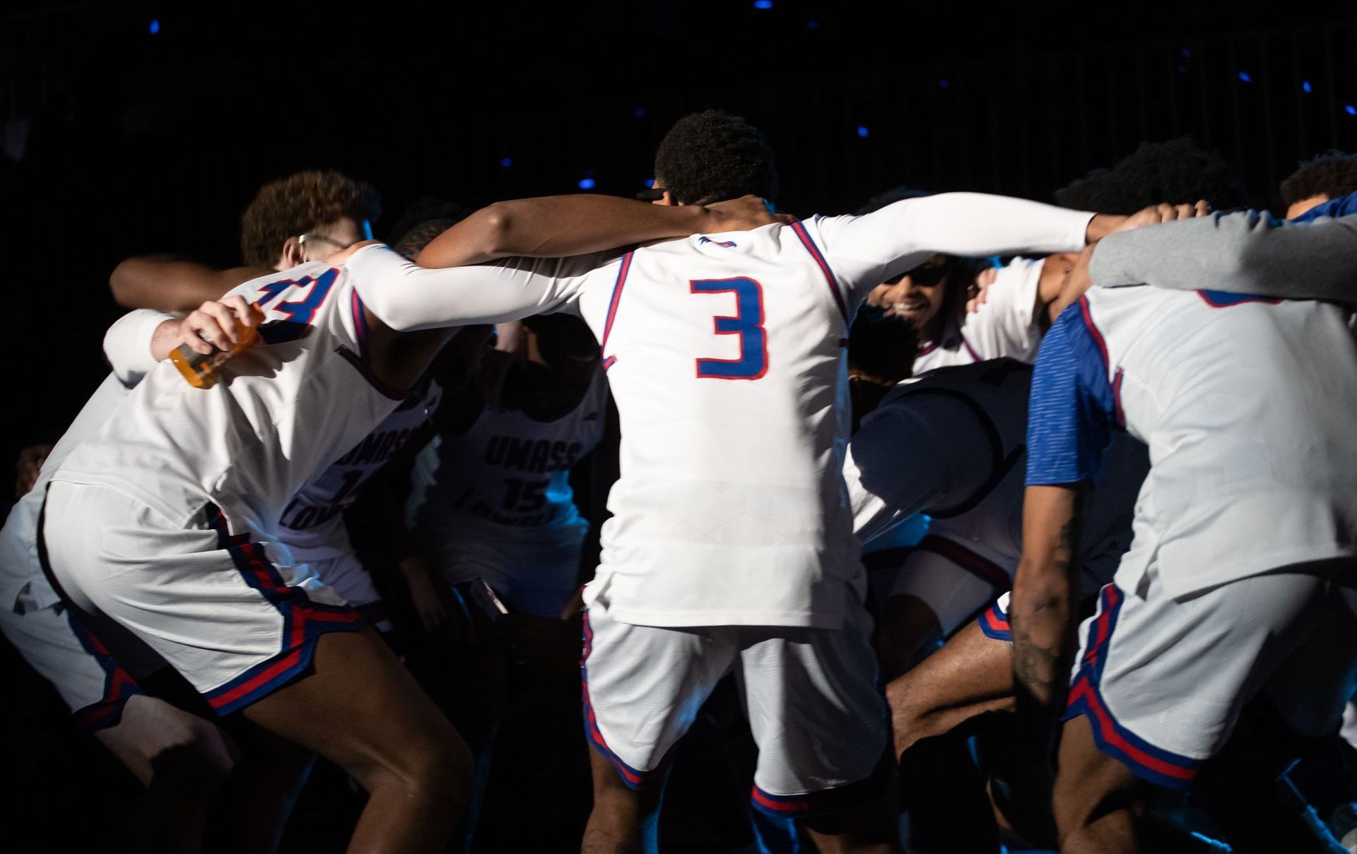 Basketball team huddled together, arms around each other, wearing white uniforms with blue accents.