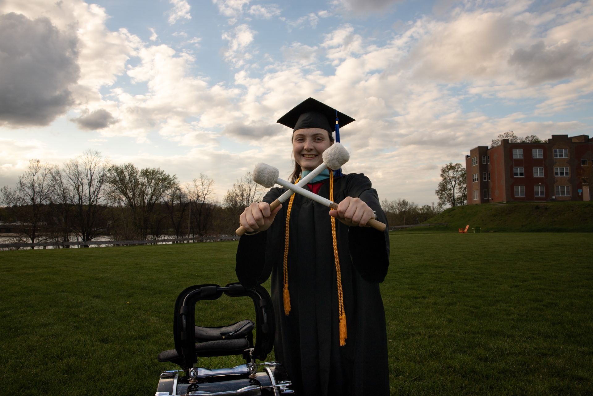 Person in graduation gown holding drum mallets, smiling outdoors near a drum set.