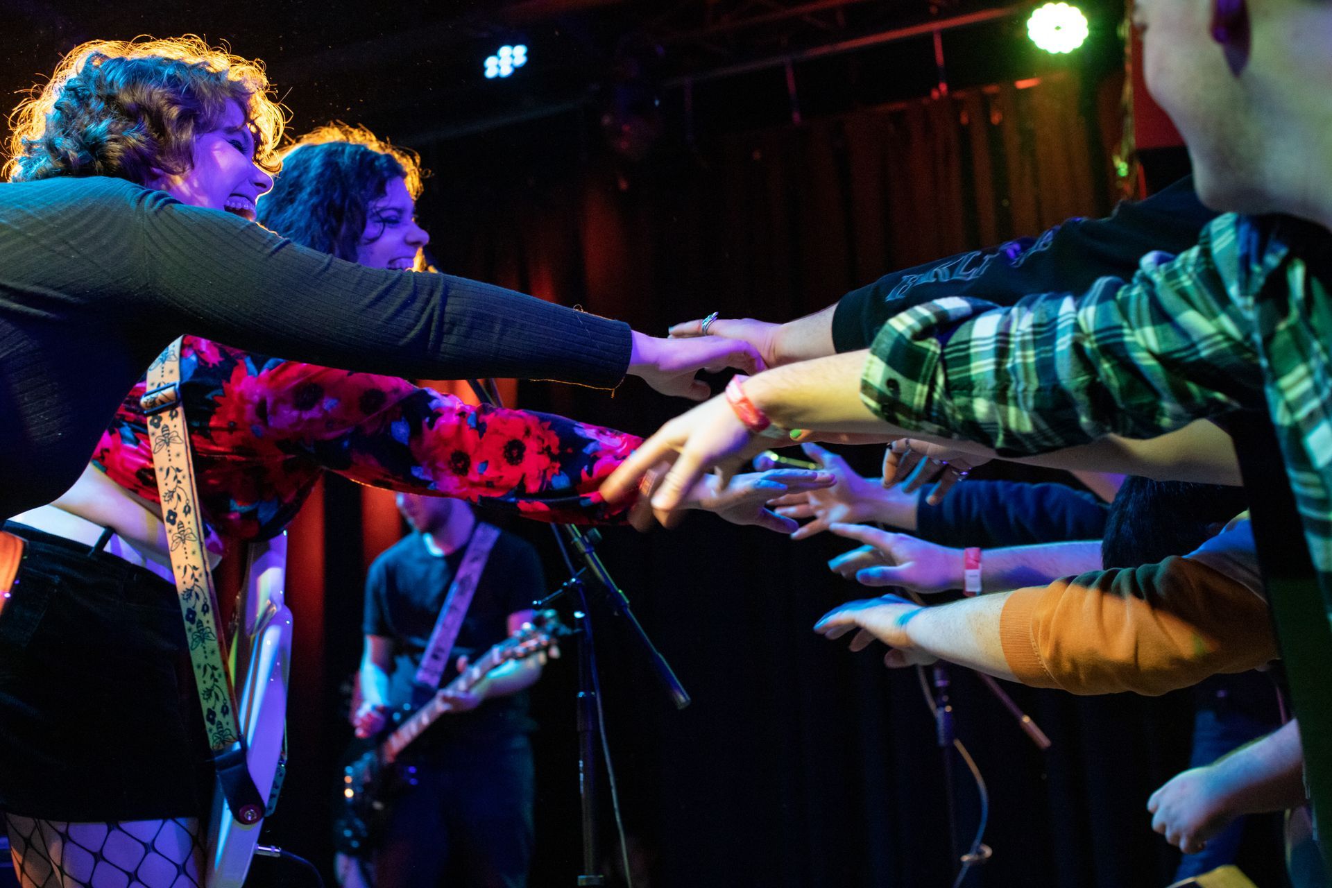 Band reaching out to touch hands with audience in a dimly lit concert venue. Stage lights illuminate the scene.