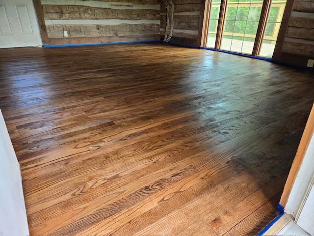Refinished hardwood floor in a room with log walls and large windows, reflecting light.
