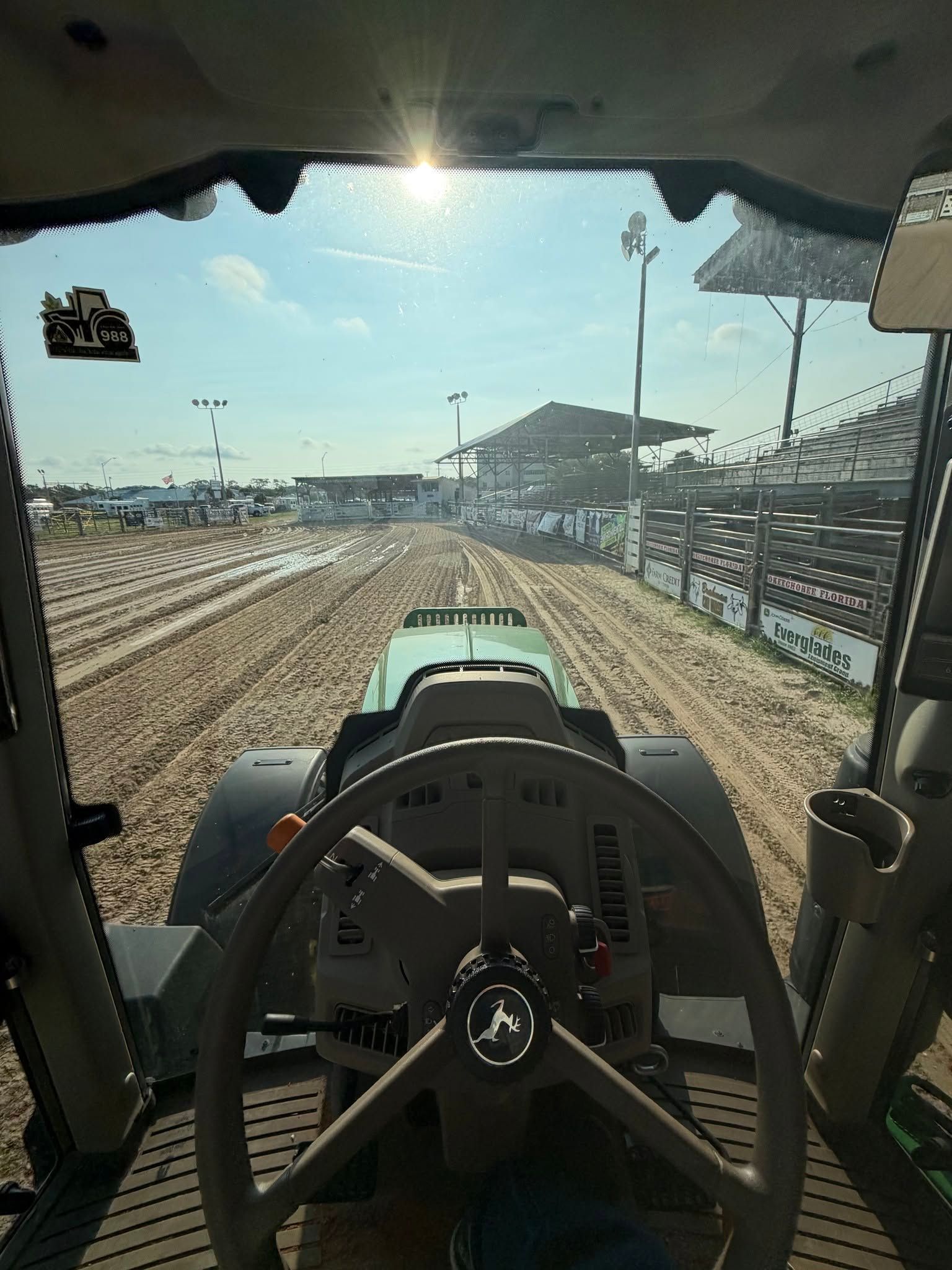 View from inside a tractor cab driving on a sandy track at an outdoor event, with bleachers ahead.