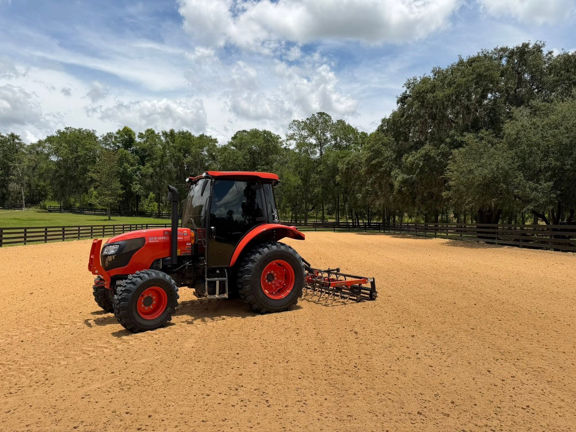 Red tractor with mower attachment in a dirt arena beside trees and fencing under a cloudy sky