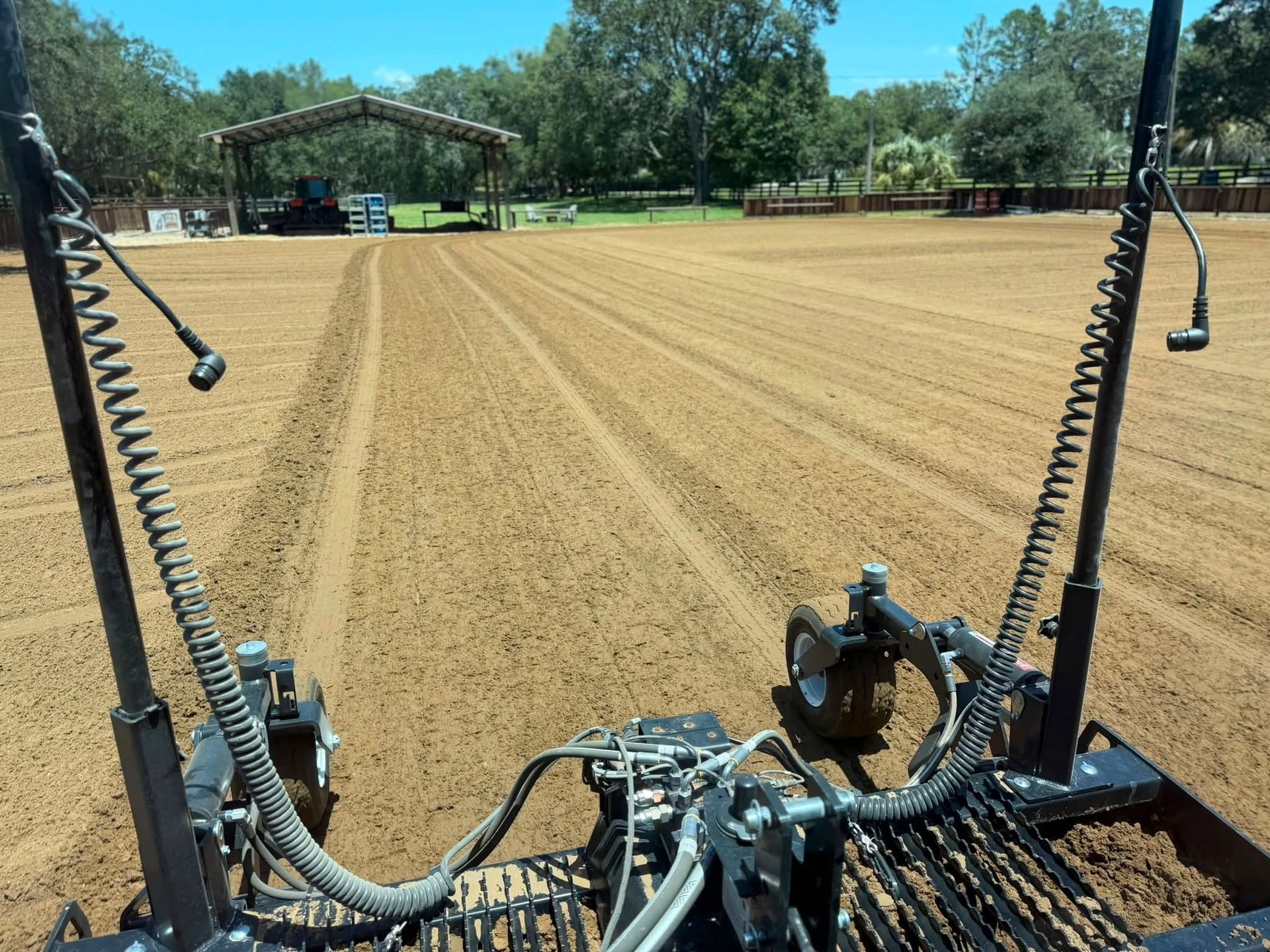 View from a tractor over a freshly groomed dirt arena with a covered riding area and trees in the background
