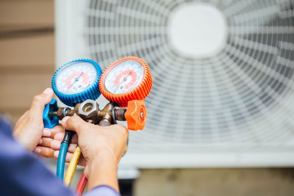 Hands holding HVAC manifold gauges with blue, red, and orange hoses in front of an outdoor fan unit
