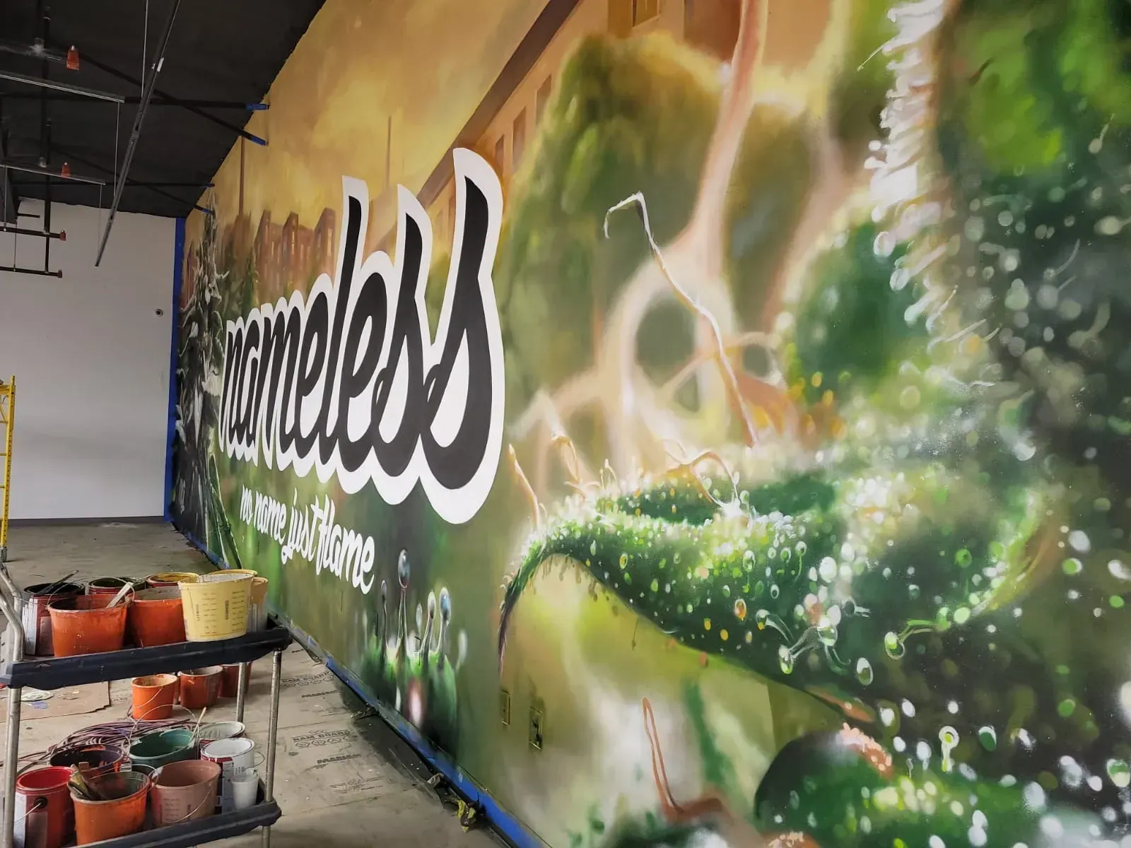 Graffiti mural with the word “homeless” in white on a green wall, beside buckets and supplies indoors