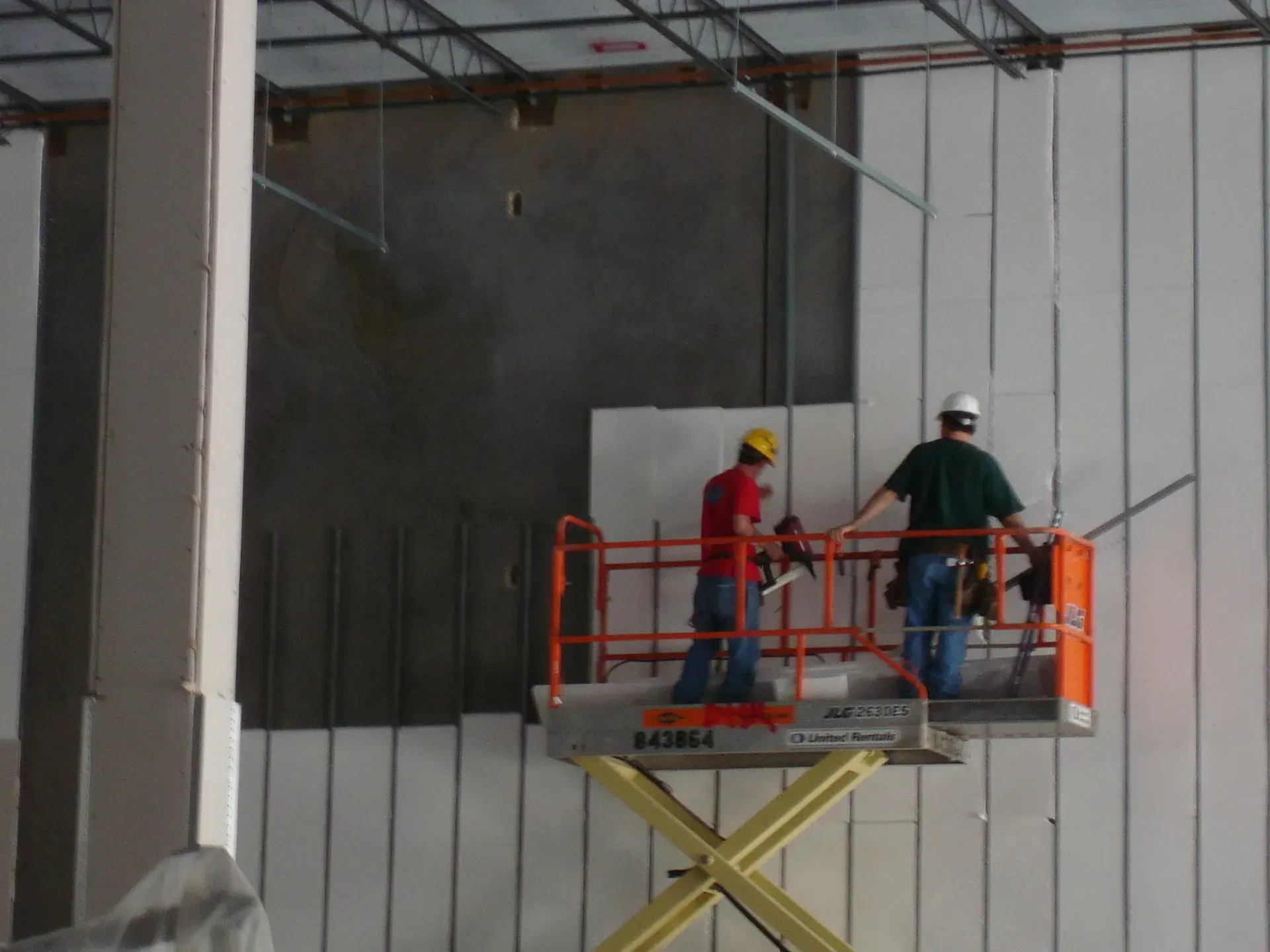 Two workers on an orange scissor lift installing panels on a building wall