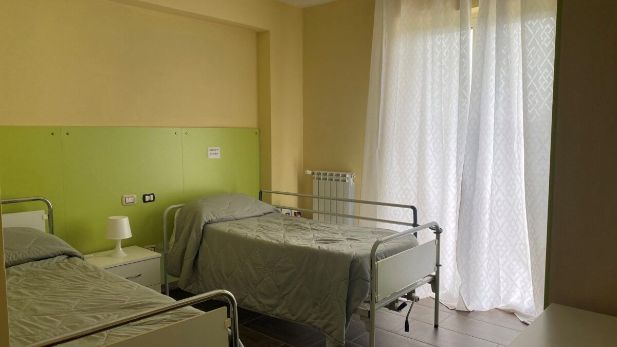 Due letti in una stanza con pareti verdi e gialle, una finestra con tende bianche e un comodino.