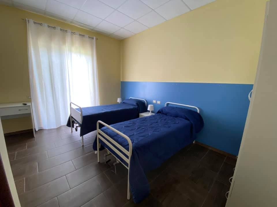 Due letti singoli in una stanza con pareti blu e gialle, una finestra e pavimento in piastrelle scure.