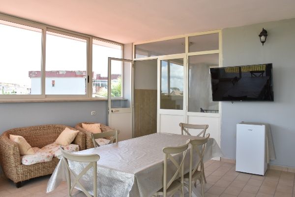 Veranda con divano in vimini, tavolo, sedie, TV e minifrigo vicino alle finestre e a una porta.