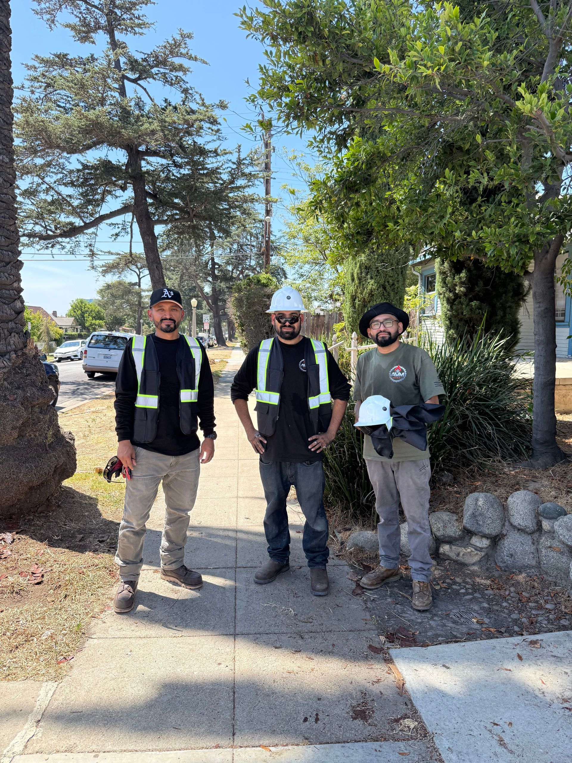 Three people standing on a sidewalk in a sunny park, wearing dark vests and hats.