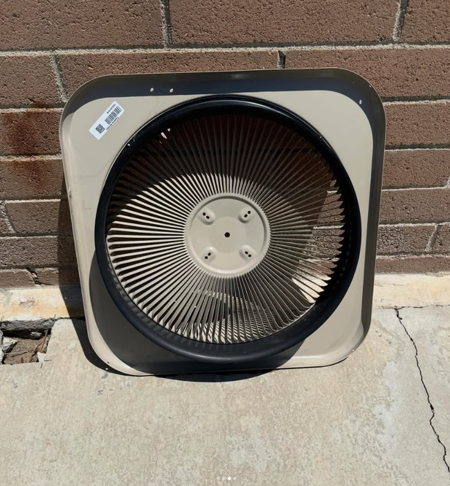 Box fan with a square beige frame leaning against a brick wall on a concrete surface