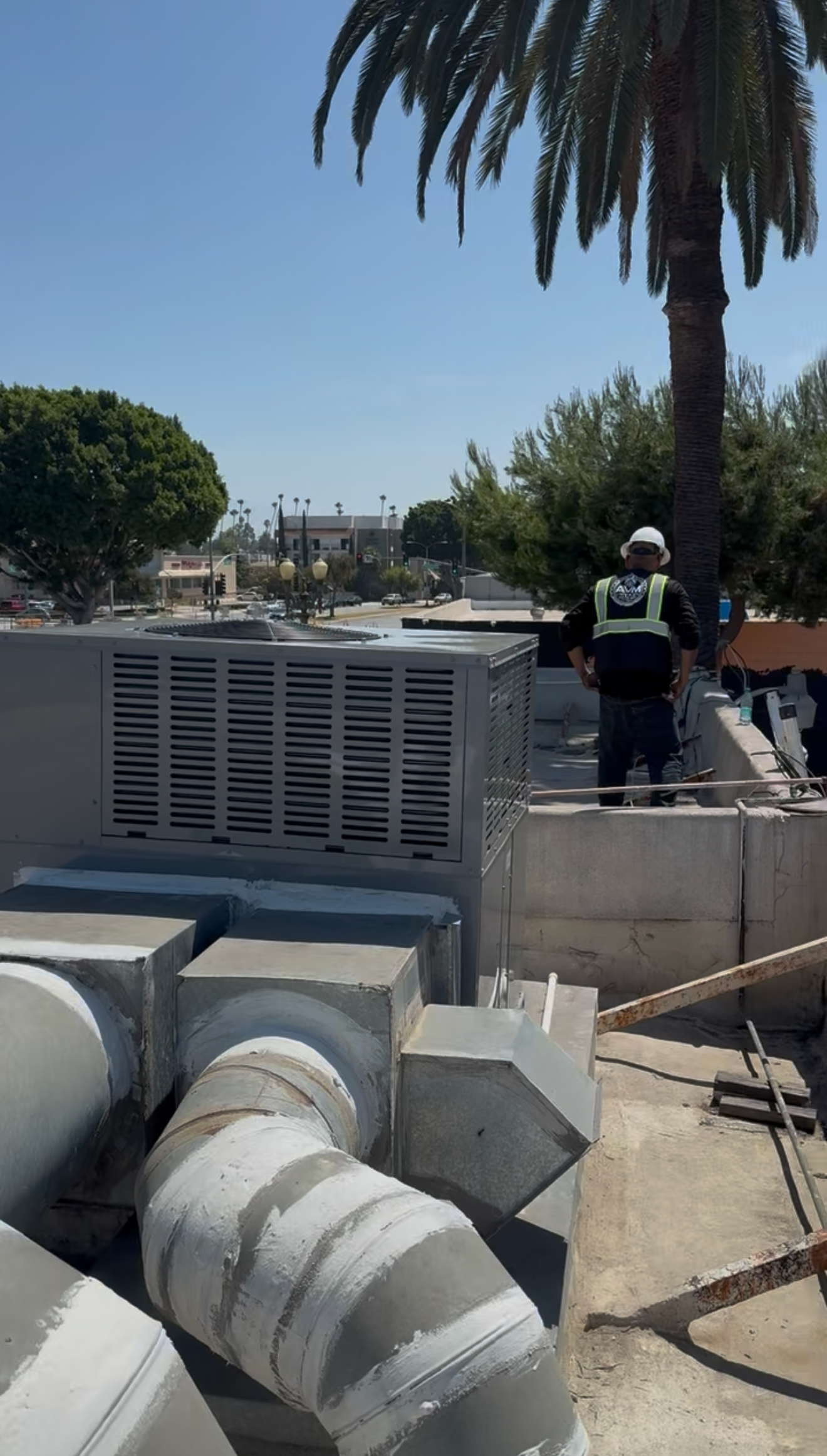 Outdoor utility area with large metal HVAC units, pipes, and a worker standing near a palm tree