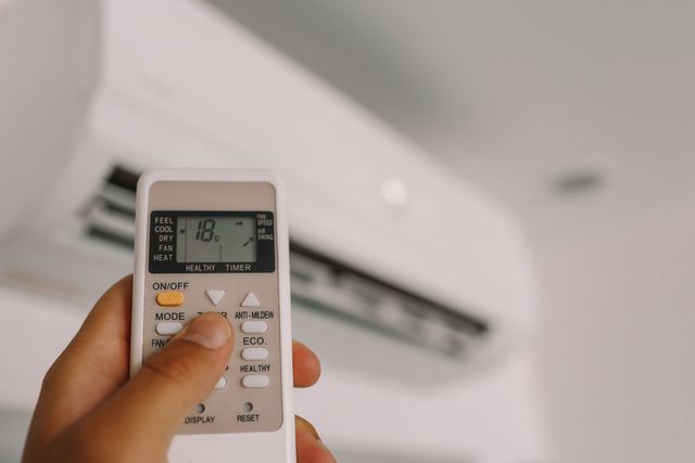Hand holding a remote aimed at a wall-mounted air conditioner in a room