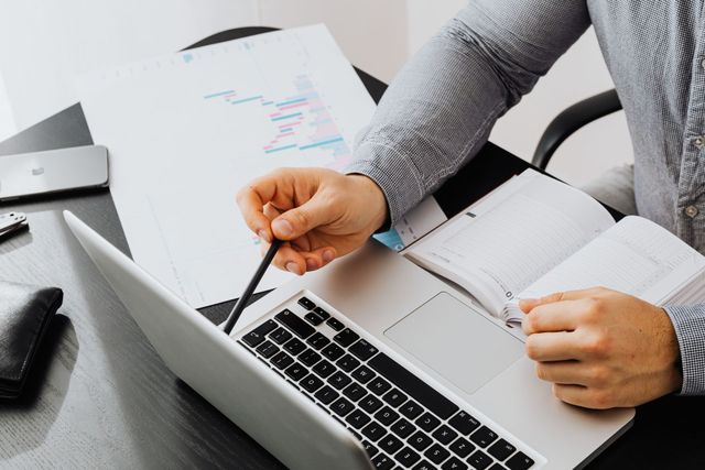 Person pointing at a laptop keyboard beside printed charts and a notebook on a desk