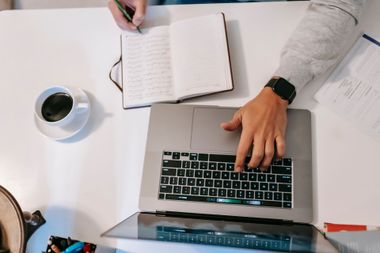 Overhead view of hands writing in a notebook beside a laptop and coffee on a white desk