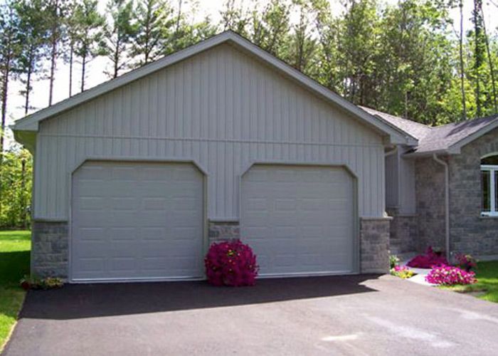 A light-colored two-car garage with vertical siding and stone accents, flanked by a stone house and green trees.