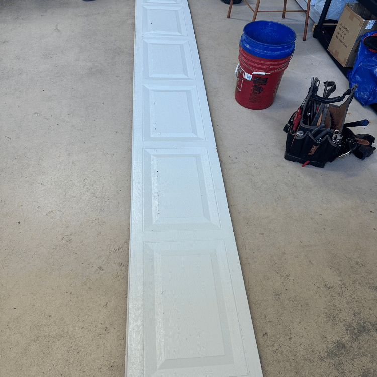 A white garage door panel lies on a concrete floor next to a red bucket and a black tool bag in a workspace.