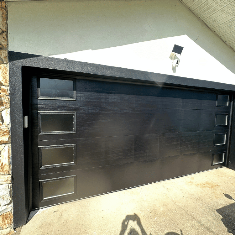 A modern black garage door with rectangular windows on both sides, installed in a white house exterior.