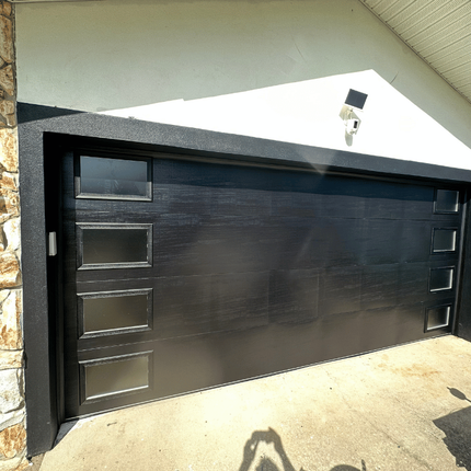 A modern black garage door with rectangular windows on both sides, installed in a white house exterior.