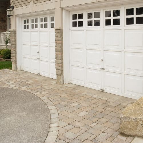 White garage doors with windows above a paver driveway, next to a stone-textured house exterior.