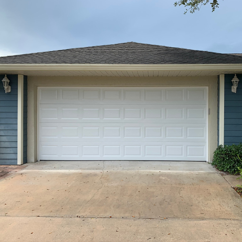 A beige house exterior featuring a white paneled garage door flanked by blue siding and wall-mounted outdoor lights.
