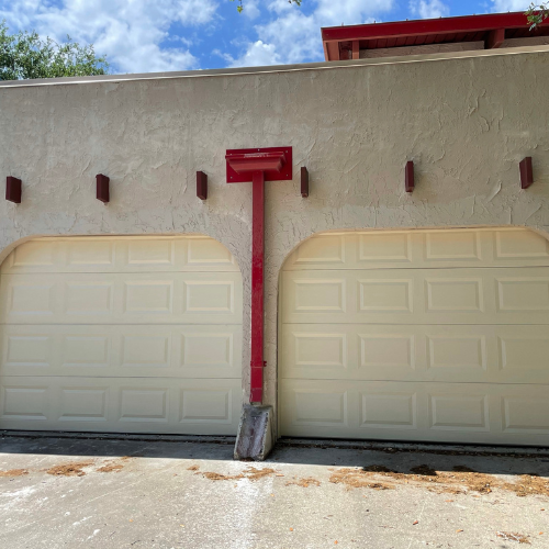 A two-car garage with cream-colored doors and a red central drainpipe on a textured beige stucco wall.