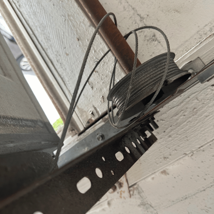 A garage door cable has come off its drum and is hanging loosely beside the metal track and mounting hardware.