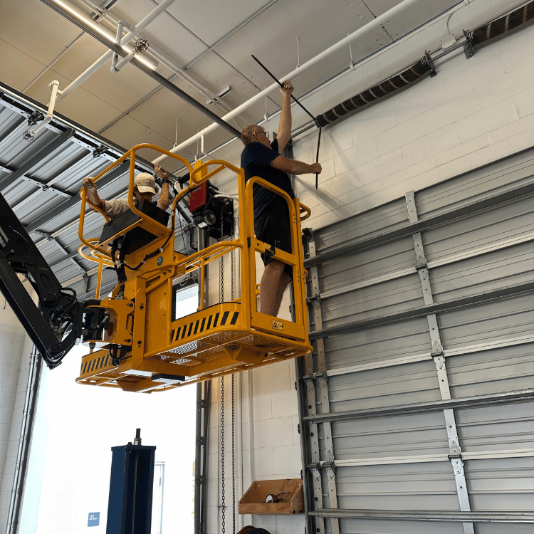 Two workers in a yellow aerial lift platform work on a metal garage door track mounted to a white ceiling.