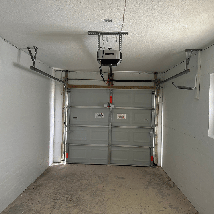 An interior view of a gray residential garage with a closed door, ceiling-mounted opener, and bare concrete floor.