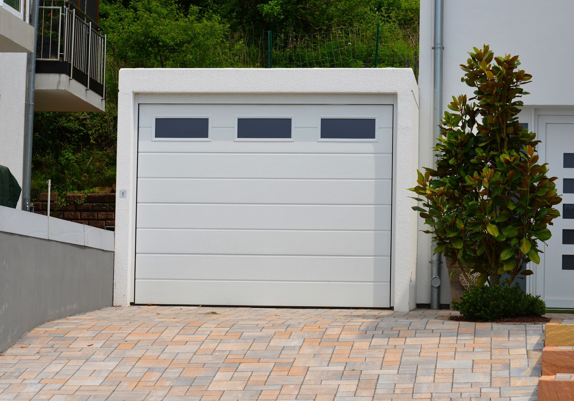 White garage door with three small rectangular windows, set in a white wall above a stone driveway.