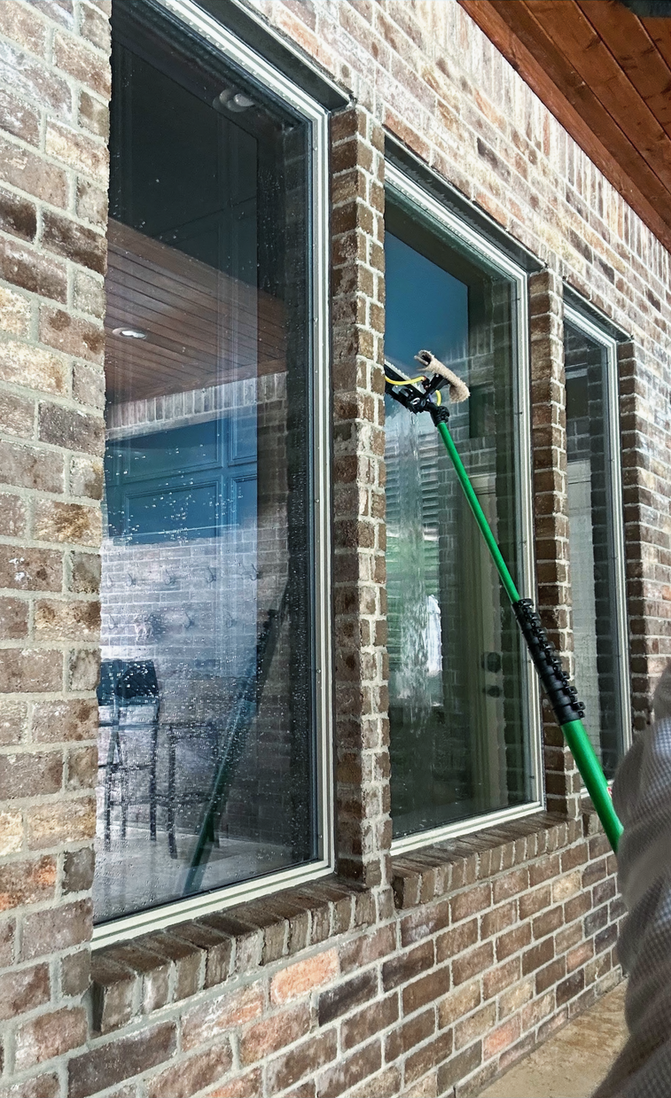 A person on a ladder applies a protective film to a large glass window on an outdoor patio.