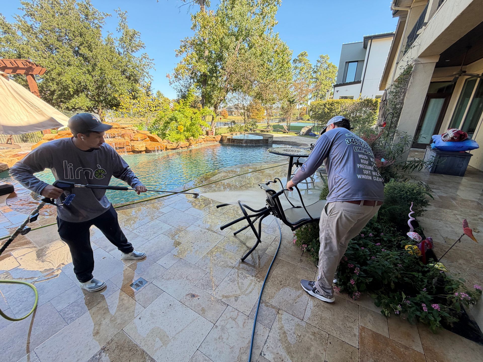 Two people pressure-wash a stone patio beside a house, spraying water toward a garden