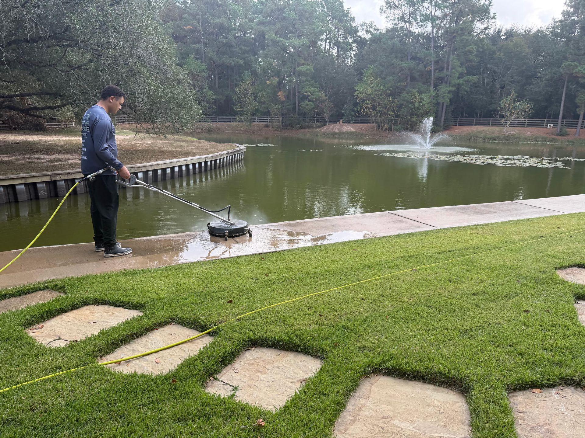 A person power-washes a concrete walkway beside a lake with a fountain and green lawn.