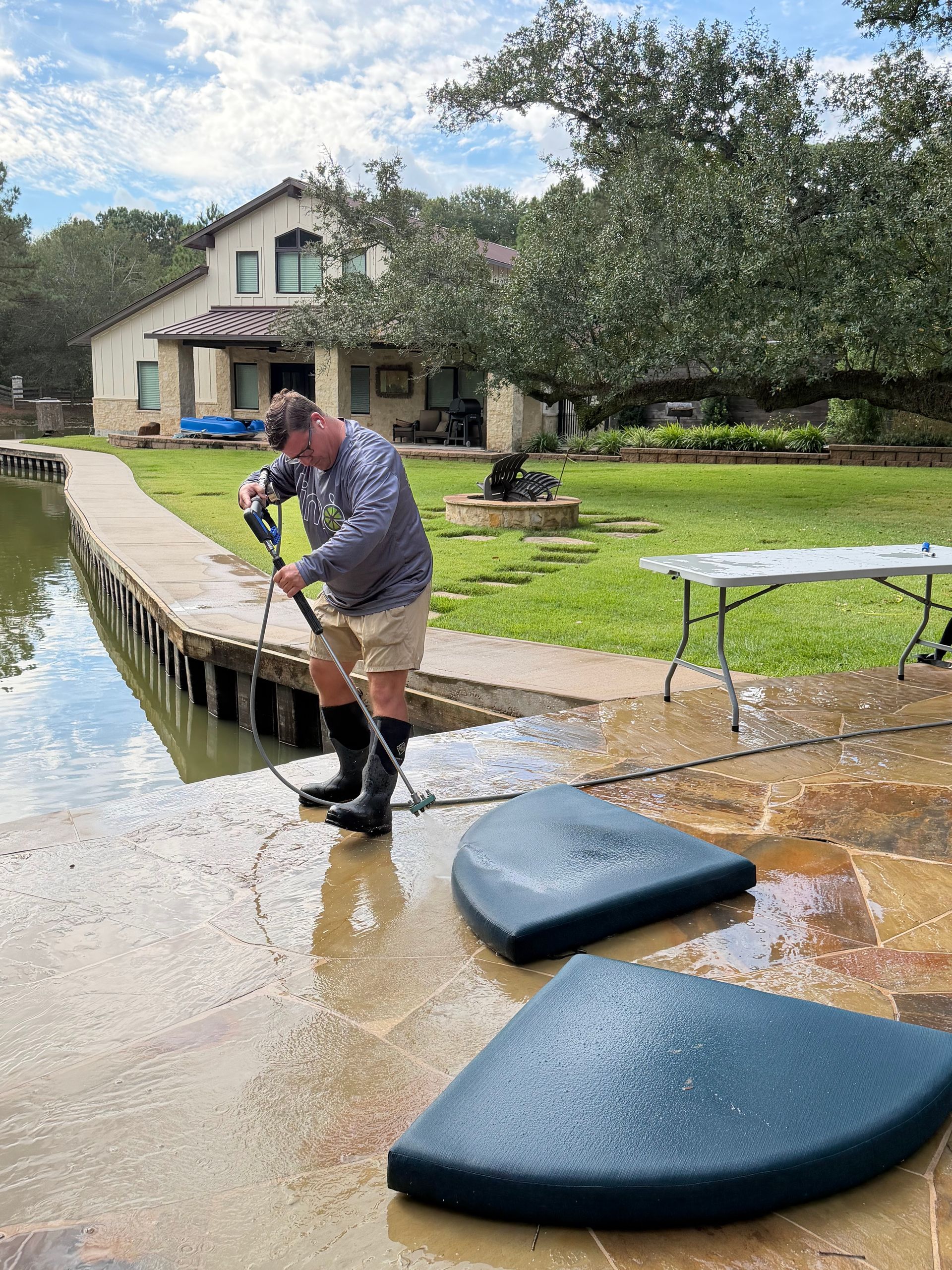 A person in boots pressure-washes a stone patio by a pond in front of a house, with two rounded dark mats on the ground.